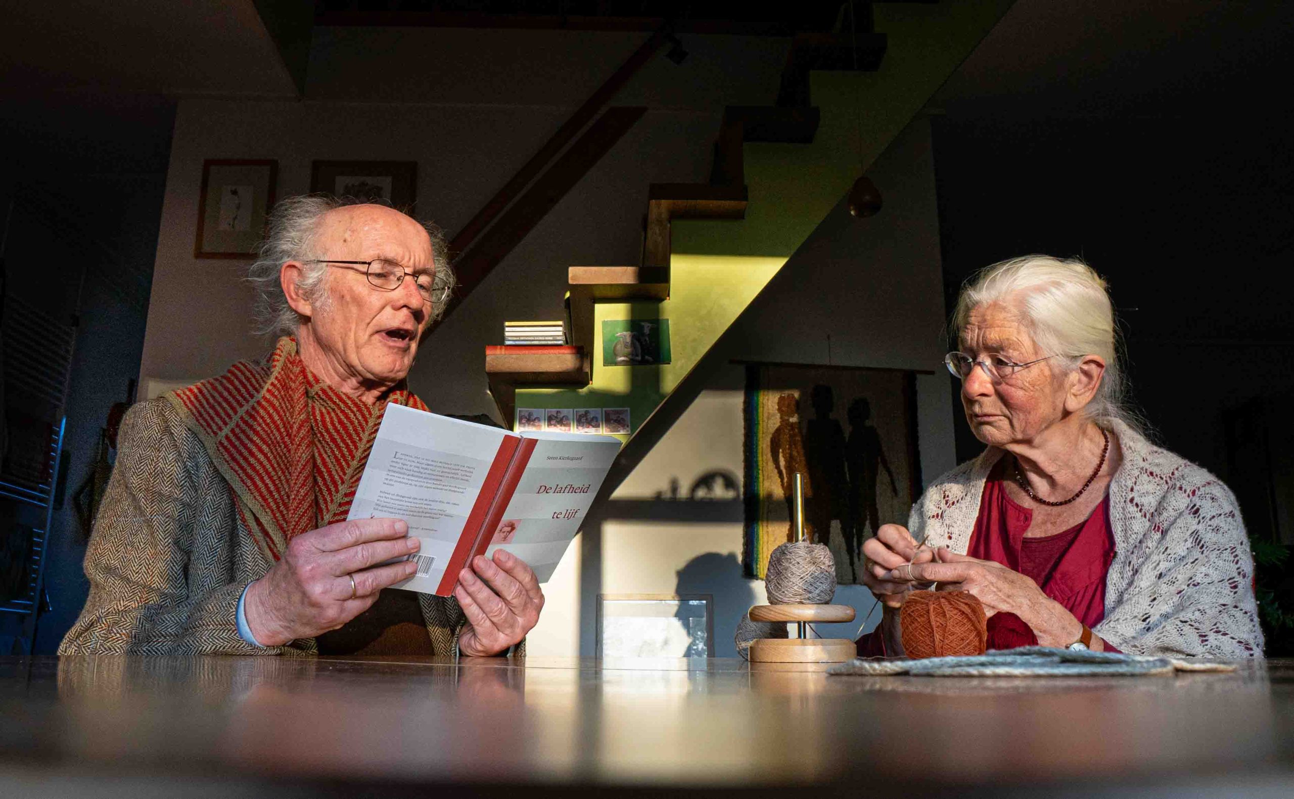 Kierkegaardvertalers Andries Visser en Lineke Buijs. Op de foto is Andries te zien die 'De lafheid te lijf' van Kierkegaard voorleest aan zijn vrouw Lineke, die bezig is met haar breiwerk.